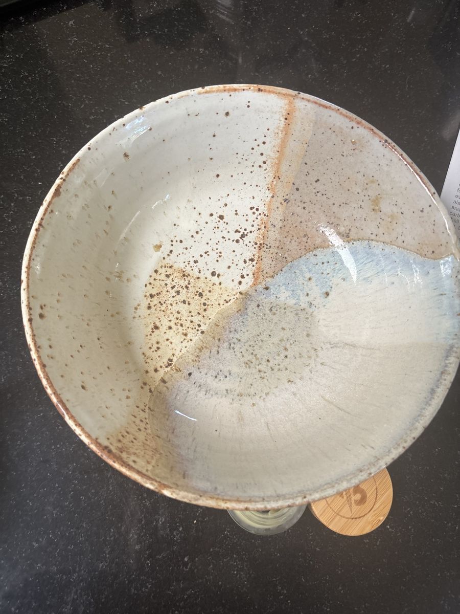 The speckled bowl from above on a dark counter — cream glaze with brown speckle and a sweep of pale blue